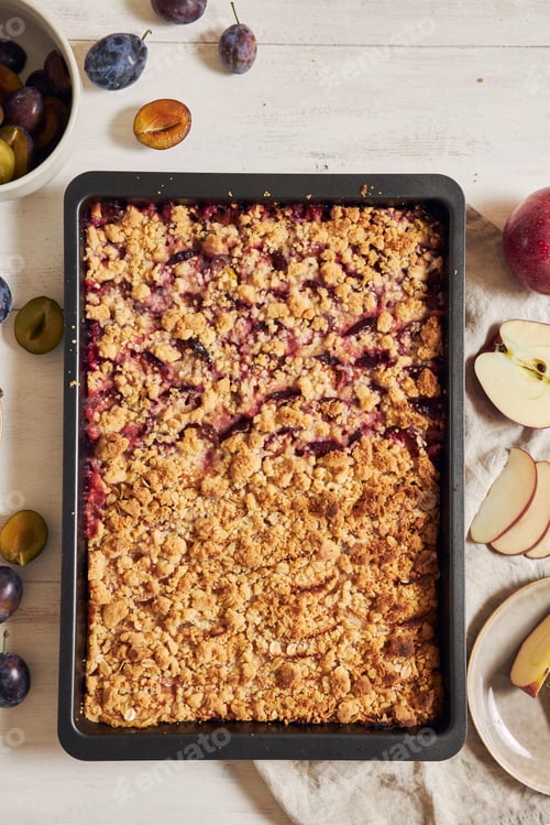 Preview: Top view of freshly baked plum and apple cake in a pan and fresh plums and apples on a white table