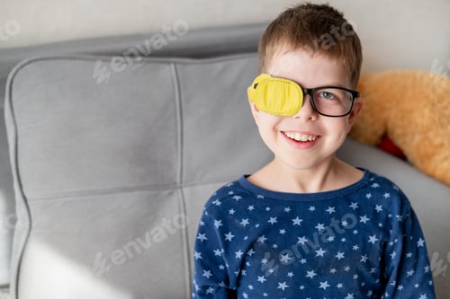 Preview: portrait of little boy wearing glasses with occluder to correct vision sitting on sofa at home
