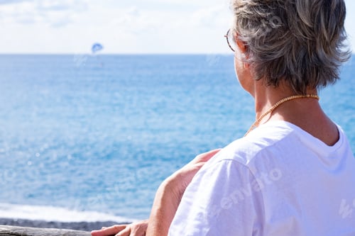 Preview: Back view of senior woman white dressed, leaning against a wooden balcony facing the sea
