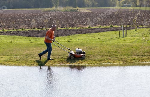 Preview: Photo of a middle-aged man mowing the grass at the pond during the beautiful spring evening.