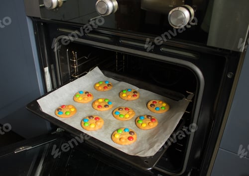 Preview: The baking tray with handmade cookies with multicolored dragees on baking paper in the oven.