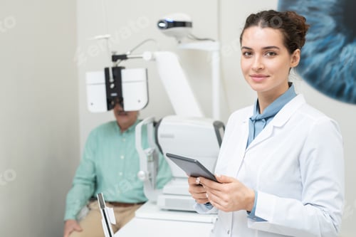 Preview: Young female ophthalmologist in whitecoat looking at you while using tablet
