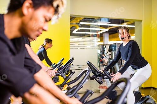 Preview: Pretty authentic female instructor with headset in fitness class exercise with group in cycling room