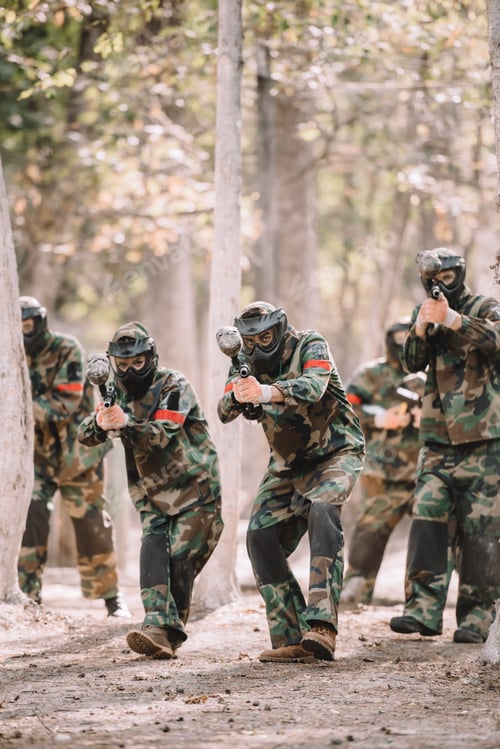 Preview: paintball team in uniform and protective masks running and aiming by paintball guns outdoors