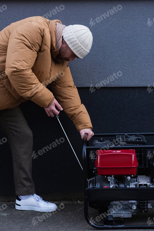 Preview: side view of man in down jacket and knitted hat starting electricity generator outdoors
