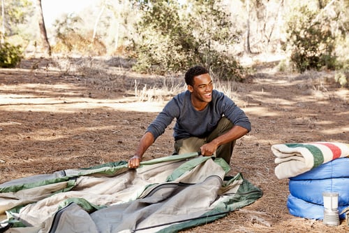 Preview: Young man kneeling in forest putting up tent