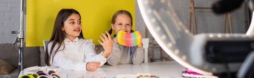 Preview: brunette blogger talking near smiling girl showing spiral toy in front of blurred circle lamp,