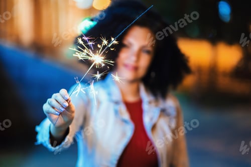 Preview: Young beautiful woman with very curly afro hair dancing with bengal fire at night illuminated street