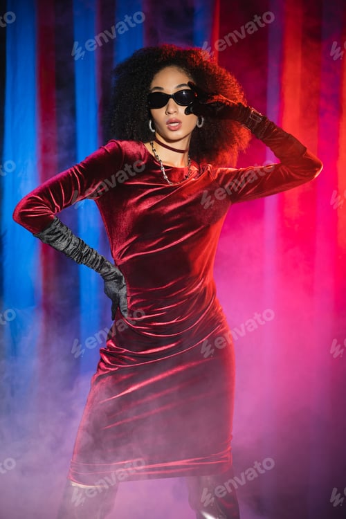 Preview: Stylish african american woman in velvet dress and sunglasses on red background with blue light