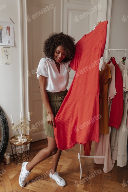 Preview: Beautiful curly young woman in khaki shorts and white blouse smiles and holds hanger with red dress