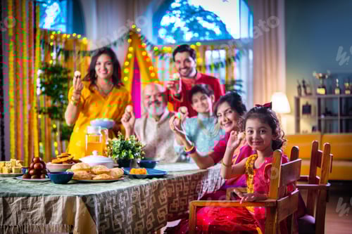 Preview: Portrait of happy Indian family in traditional outfit celebrating Diwali at home eating food
