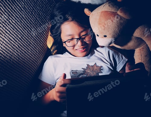 Preview: A girl and her teddy bear lying on sofa watching movie on tablet