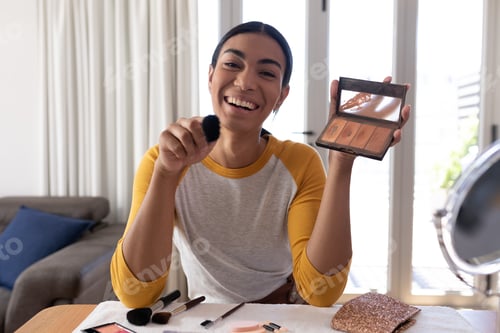 Preview: Mixed race gender fluid man making vlog holding foundation powder makeup and brush, smiling