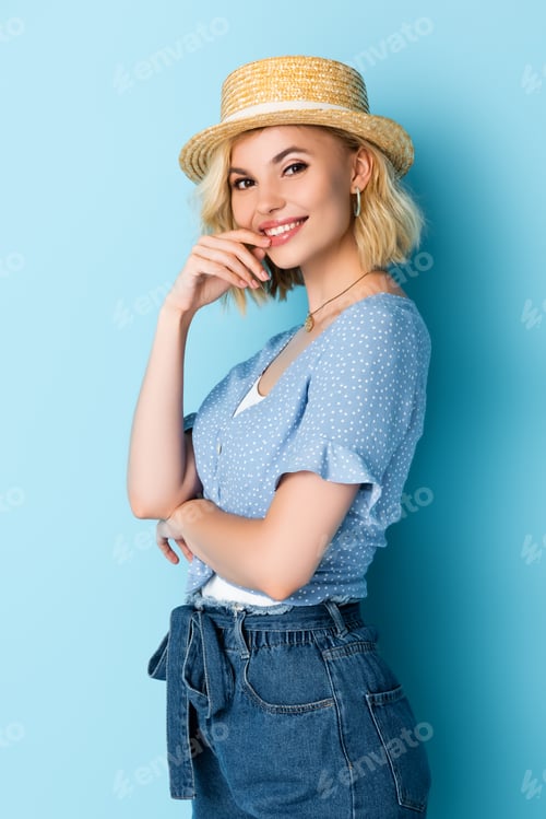 Preview: young woman in straw hat touching face and looking at camera on blue