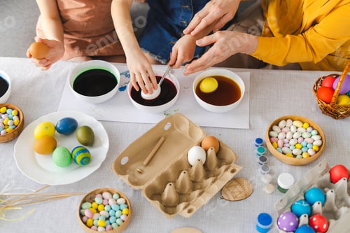 Preview: Top view grandmother helping granddaughter dyeing easter eggs at home selective focus on hands