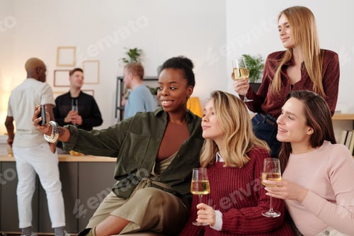 Preview: Happy young intercultural women with flutes of champagne making selfie