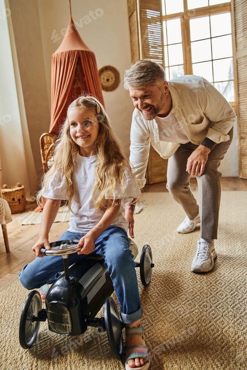 Preview: happy man assisting cheerful daughter riding toy car in modern living room at home, playing together