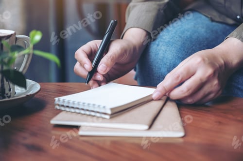 Preview: Closeup image of a woman's hand preparing to write on blank notebook with coffee cup on wooden table