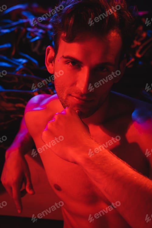 Preview: young shirtless man looking at camera while posing near black blurred bedding in red lighting