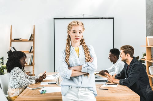 Preview: portrait of upset businesswoman with arms crossed looking at camera and colleagues working behind in