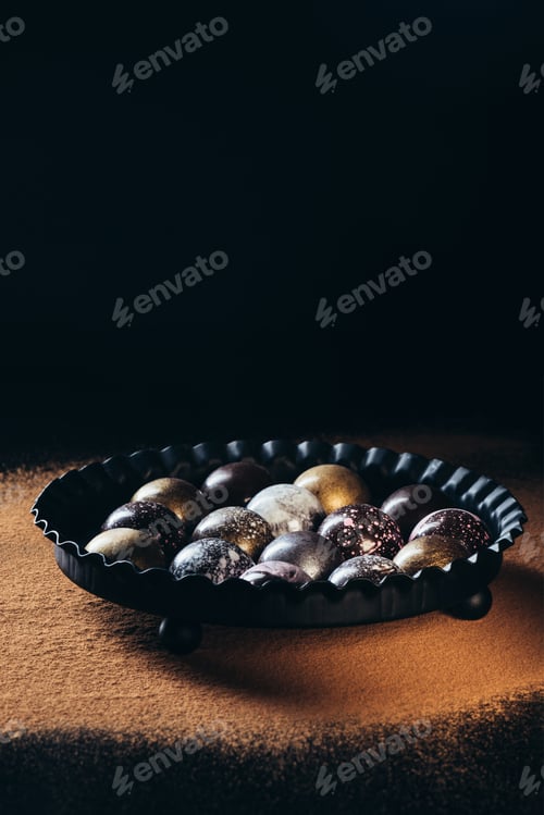 Preview: pile of different chocolate candies in bowl on surface with cocoa powder