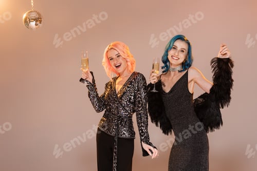 Preview: excited and stylish nonbinary friends smiling at camera and posing with champagne glasses on beige