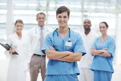 Preview: Young male doctor standing smiling with his arms folded and his colleagues in the background