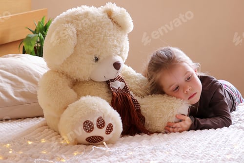Preview: A little pretty girl looks with a bored look. A teddy bear sits nearby. Bored mood.