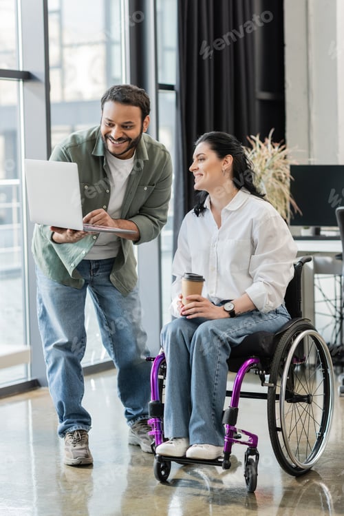 Preview: cheerful indian businessman showing startup project on laptop to disabled woman in wheelchair