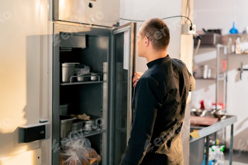 Preview: chef stands in kitchen in a black uniform and looks through what is on the list in the refrigerator