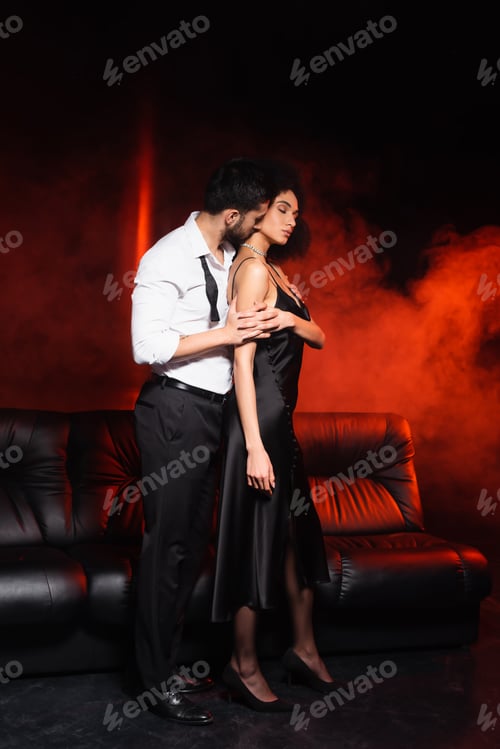 Preview: Man in formal wear kissing neck of african american girlfriend on black background with red lighting