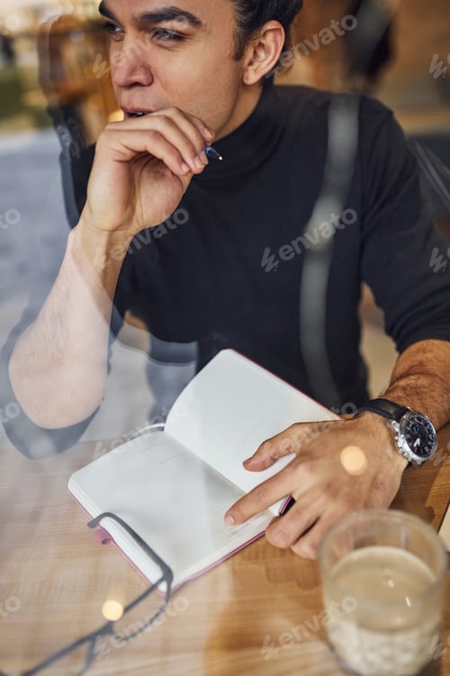 Preview: Young male writer with black curly hair sitting indoors in cafe with pen and notepad