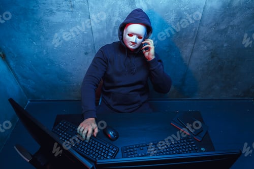 Preview: Anonymous hacker wearing face mask working on computer in dark room