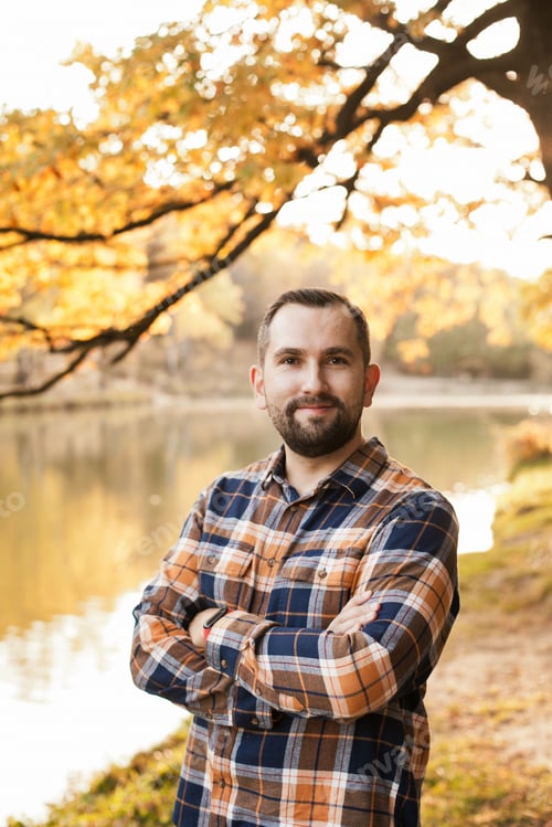 Preview: a guy in checkered shirt with beard smiling, standing alone in autumn park by the lake near tree