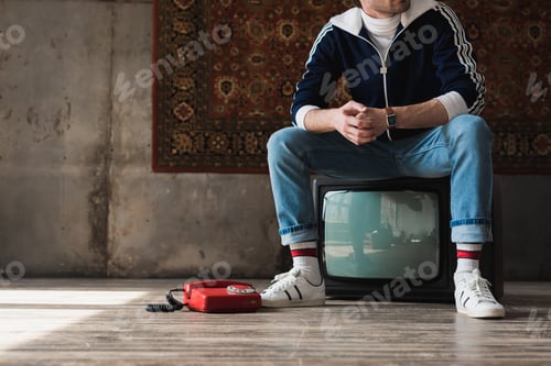 Preview: handsome young man in vintage clothes sitting on retro tv set near red wired telephone in front of
