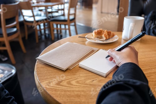 Preview: Closeup image of a hand writing on blank notebook with coffee cup and croissant on table in cafe