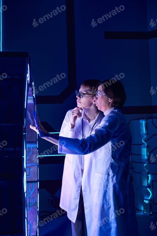 Preview: advanced learning, future, middle aged woman scientist showing innovative device to young intern