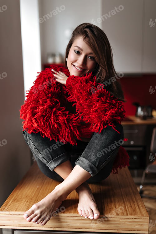 Preview: Lovely woman in black jeans and red sweater sits on kitchen table and smiles sweetly
