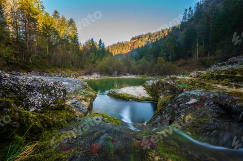 Preview: Tranquil mountain stream meandering through a lush green forest, featuring jagged rocky terrain