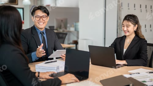 Preview: Smiling businessman holding tablet talking to woman coworker while sitting at wooden table in office