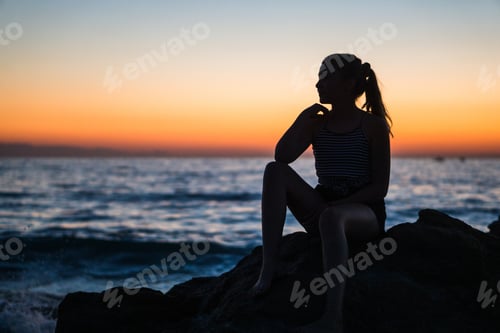 Preview: Silhouette of a young female sitting on a rock by the ocean staring into the breathtaking sunset