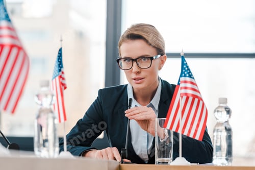 Preview: selective focus of beautiful diplomat in eyeglasses touching microphone near american flags