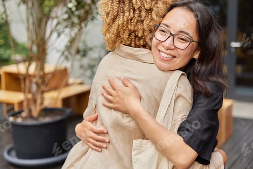 Preview: Happy Asian woman in spectacles greets female friend and embrace each other dressed casually pose ou
