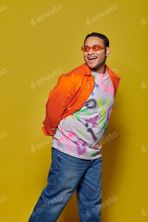 Preview: amazed indian man in sunglasses and vibrant attire looking away and smiling on yellow background