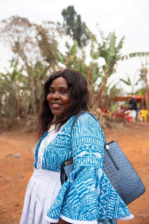 Preview: A smiling African woman dressed in traditional clothing and carrying a bag, vertical photo.