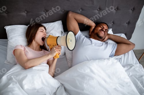 Preview: Young Caucasian woman shouting into megaphone at her sleeping husband, trying to wake him up in