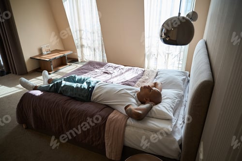 Preview: Thoughtful african american man lying and resting on bed in hotel room at day