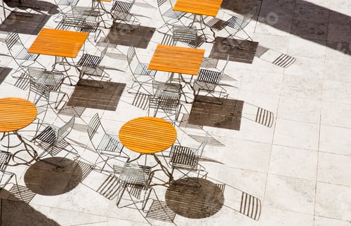 Preview: High angle view of wooden tables and metal folding chairs casting shadows onto brown paving.