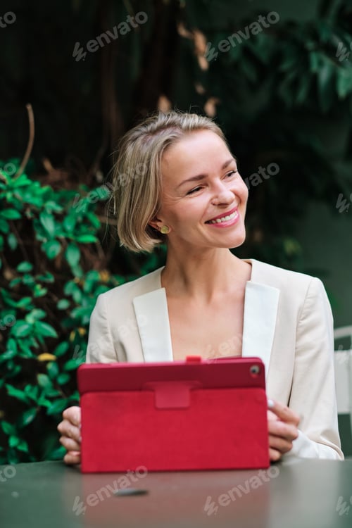 Preview: Woman looking away and smiling while using a digital tablet sitting on a table outdoors.