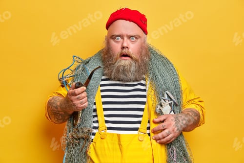 Preview: Plump bearded professional man mariner stares at camera with displeased shocked expression holds smo